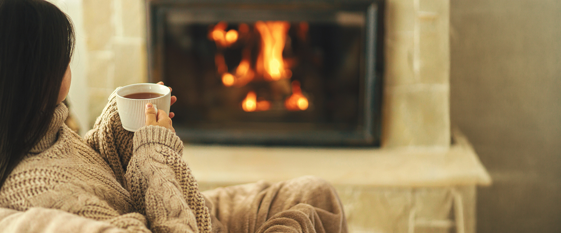 Persoon zit onder een warme plaid met een mok thee of koffie voor een brandende open haard.