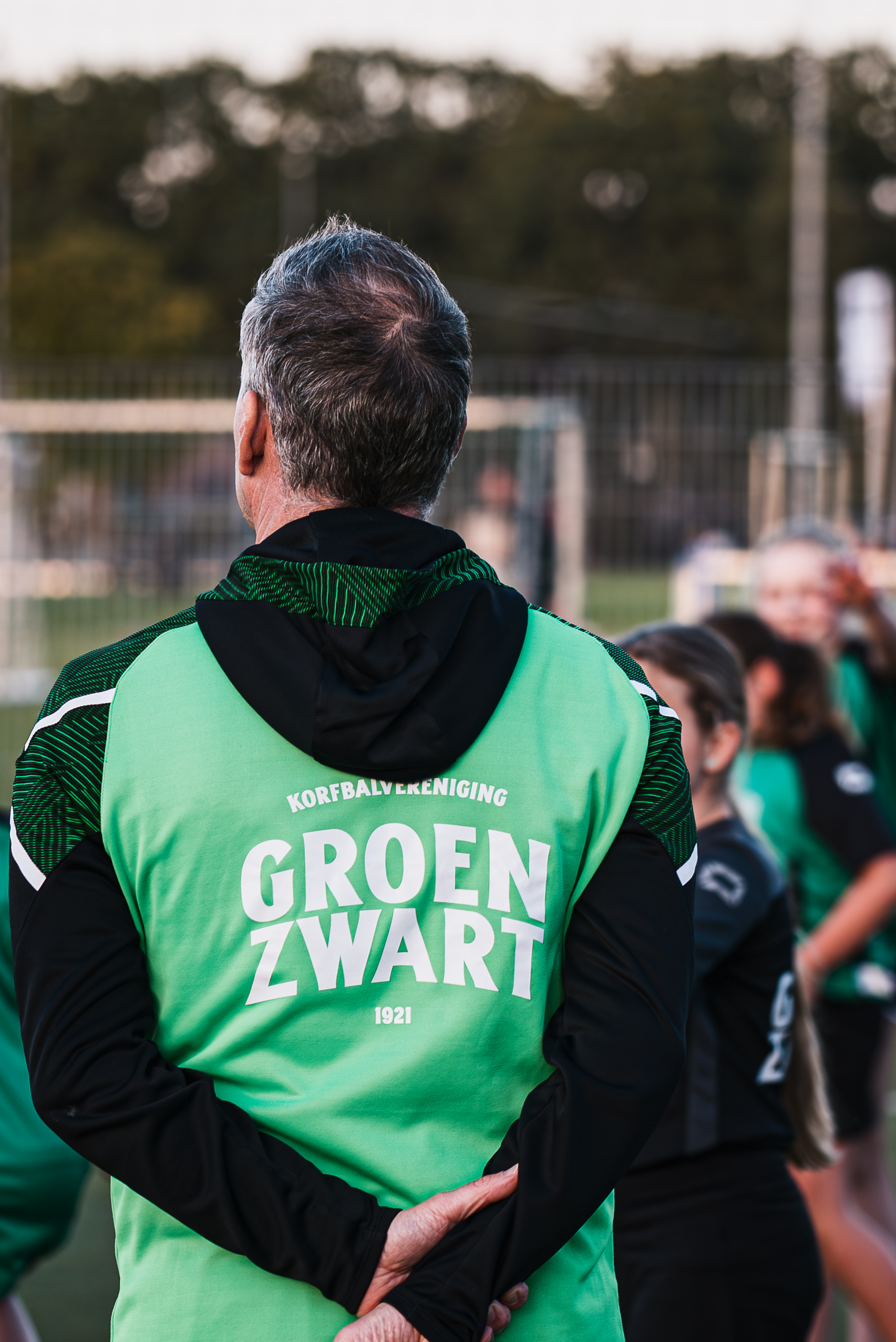 Persoon in Groen Zwart-clubkleding op het korfbalveld, met andere clubleden op de achtergrond.